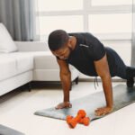 Homem fazendo flexão de braços durante treino em casa, utilizando tapete e acompanhando exercício por notebook.